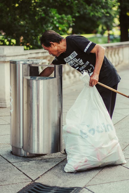 An elderly woman with short dark hair, wearing a black T-shirt with white text and dark trousers, is seen disposing of rubbish into a public metallic trash bin located on a paved sidewalk. She is bent forward, reaching into the receptacle to deposit waste, holding a large white plastic bag filled with trash in her left hand, which is supported by her hip. The trash bin has a shiny stainless steel finish and is situated near a stone railing, with lush green trees and foliage in the background indicating an outdoor park or promenade setting. The scene appears to be well-lit by natural daylight, and the surroundings are clean and orderly, reflecting efforts of responsible waste disposal. This image illustrates an on-site, private approach to rubbish removal, aligning with alternative waste handling methods supervised by specialist services like Rubbish Removal Bayswater to support community cleanliness and environmental responsibility.