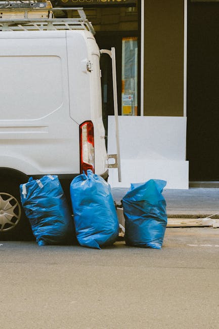 Four large blue rubbish bags filled with waste are placed on the pavement beside the rear of a white delivery van, which is parked in front of a building with large glass windows and a black exterior wall. The van's rear door is slightly open, and a white panel or board leans against the building's entrance. The bags appear to be made of durable plastic with a shiny finish, and they are slightly wrinkled and piled on one another. The scene is set outdoors in an urban environment, with visible concrete pavement and the edge of the road. The natural lighting is diffuse, suggesting an overcast sky, and the overall context relates to onsite or private waste disposal, as provided by rubbish removal services such as Rubbish Removal Bayswater, illustrating a typical scene of waste collection or clearance outside a commercial or residential property.