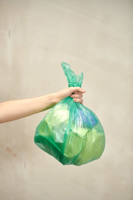 A human arm extending from the left side of the image holds a green and blue plastic bag filled with waste. The bag appears to contain soft, lightweight materials, possibly packaging or household rubbish, and is tied at the top with a twist tie or knot. The background is a plain, light beige wall, providing a neutral setting that emphasizes the plastic bag. The scene reflects an act of waste collection or disposal, consistent with rubbish removal activities provided by services such as Rubbish Removal Bayswater. The lighting is evenly distributed, highlighting the translucent quality of the plastic bag and the textures of its contents. The image conveys the concept of private or independent waste handling, relevant to rubbish clearance for flats or similar residential settings, and aligns with themes of rubbish management or disposal services in urban environments.