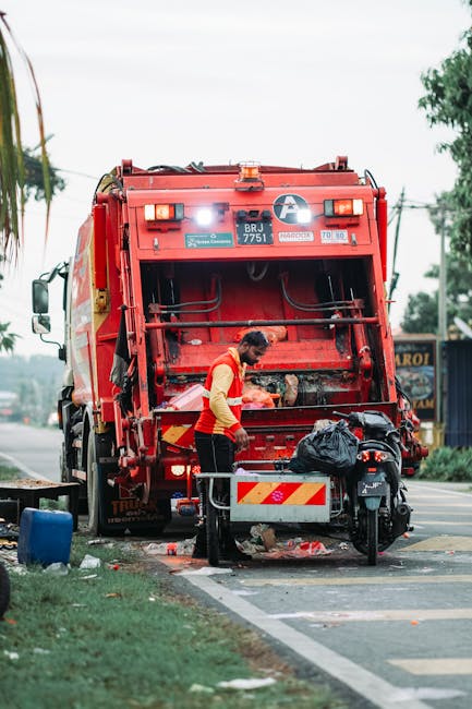 A white industrial rubbish collection truck is parked on a narrow cobblestone street in an urban area, with its rear compactor open and engaged in unloading waste. A worker dressed in a high-visibility orange vest and blue uniform is positioned on the right, holding a blue wheeled rubbish bin, preparing to empty it into the truck’s hopper. The street is lined with multi-storey buildings featuring varied exterior finishes, some with visible weathering and patches of darker paint or plaster. The surrounding environment includes parked cars, with one black vehicle directly behind the rubbish truck, and signs attached to building facades indicating parking restrictions. The scene appears to be during daylight hours with natural lighting, and the overall setting reflects an everyday urban waste collection process, emphasizing private waste handling services rather than municipal collection, consistent with the services offered by Rubbish Removal Bayswater.